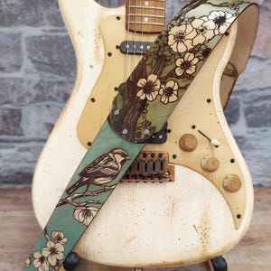 May include: A cream-colored electric guitar with a decorative leather strap. The strap features a bird perched on a branch with flowers, set against a teal background. The strap is adorned with a floral design.