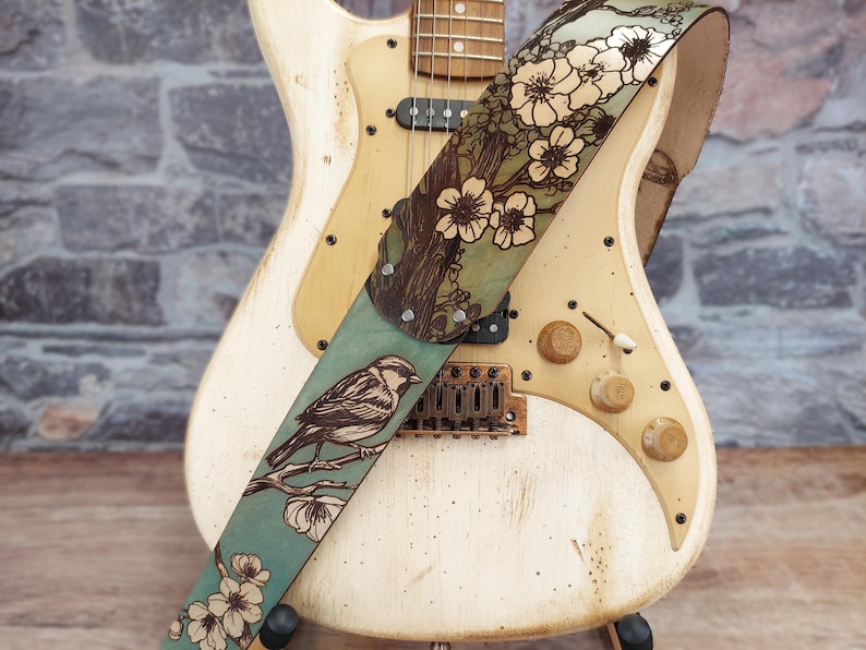May include: A cream-colored electric guitar with a decorative leather strap. The strap features a bird perched on a branch with flowers, set against a teal background. The strap is adorned with a floral design.