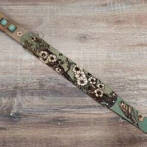 May include: A guitar strap with a floral design. The strap features a light green background with a pattern of white flowers and brown branches. The strap has a light brown leather section with green accents.