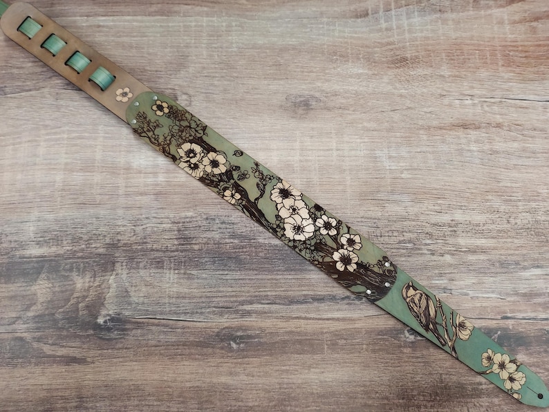 May include: A guitar strap with a floral design. The strap features a light green background with a pattern of white flowers and brown branches. The strap has a light brown leather section with green accents.