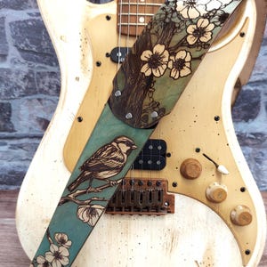May include: A cream-colored electric guitar with a decorative leather guitar strap. The strap features a detailed illustration of a bird perched on a branch with flowers, set against a teal background.