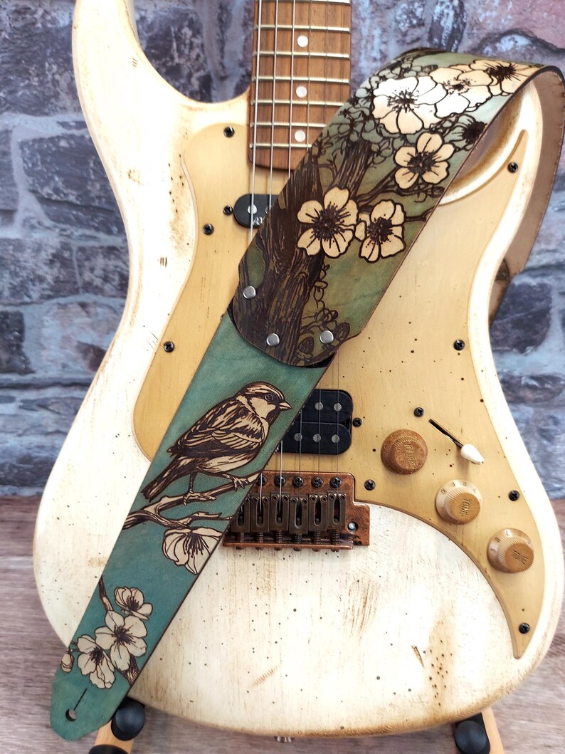 May include: A cream-colored electric guitar with a decorative leather guitar strap. The strap features a detailed illustration of a bird perched on a branch with flowers, set against a teal background.