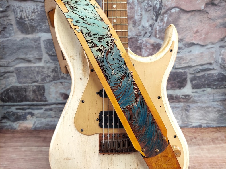 Puede incluir: Una guitarra el&eacute;ctrica color crema con una correa decorativa. La correa presenta un dise&ntilde;o de olas azules y marrones sobre un fondo beige. La guitarra tiene pastillas negras y un golpeador blanco.