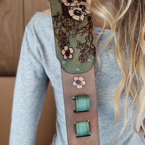 May include: A guitar strap with a floral design. The strap is made of brown leather with a green top section and teal accents. The floral design is etched into the leather and features flowers.