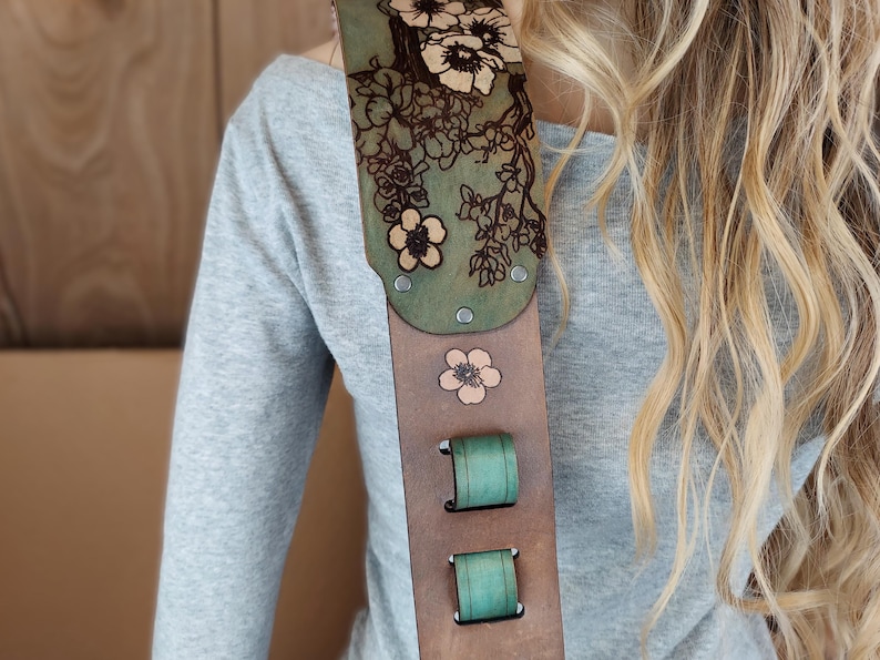 May include: A guitar strap with a floral design. The strap is made of brown leather with a green top section and teal accents. The floral design is etched into the leather and features flowers.