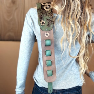 May include: A brown leather guitar strap with a floral design and teal accents. The strap features a green top section with laser-cut flowers and a series of teal-colored adjustment buckles. A single pink flower is also visible.