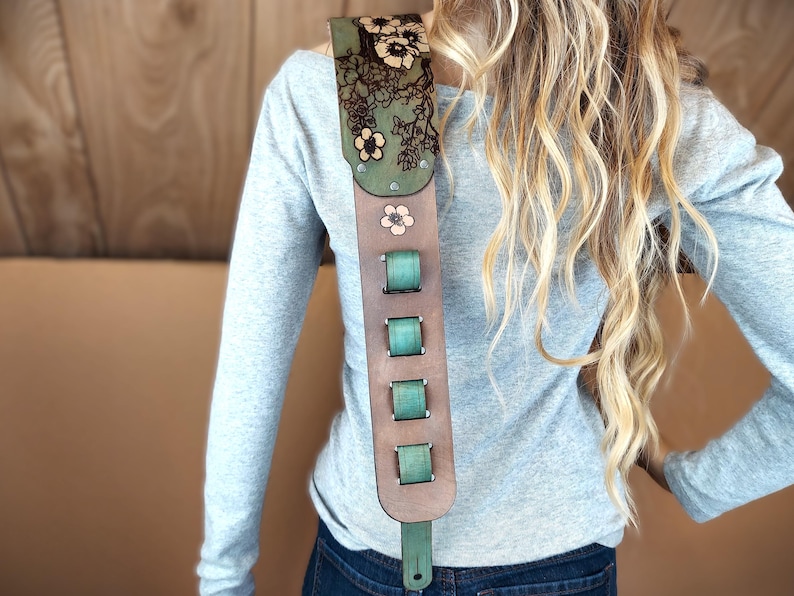 May include: A brown leather guitar strap with a floral design and teal accents. The strap features a green top section with laser-cut flowers and a series of teal-colored adjustment buckles. A single pink flower is also visible.