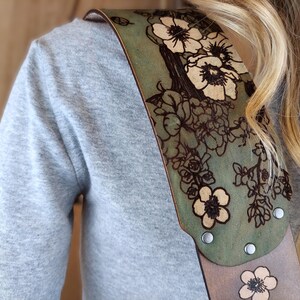 May include: A guitar strap with a floral design. The strap is made of leather and features a green and tan color scheme. The floral pattern is etched into the leather, with silver rivets securing the strap.