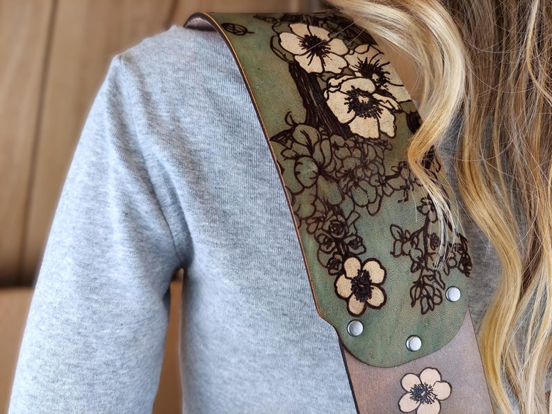 May include: A guitar strap with a floral design. The strap is made of leather and features a green and tan color scheme. The floral pattern is etched into the leather, with silver rivets securing the strap.