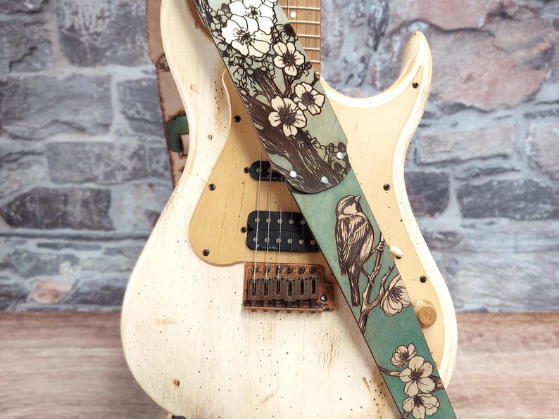 This Handmade Leather Guitar Strap features a songbird perched on cherry blossom branches against a unique hand dyed distressed finish in Muted Turquoise, Silverstone and Natural Tan with a great vintage look. The strap is adjustable on one end to fit a range of player heights and positions.