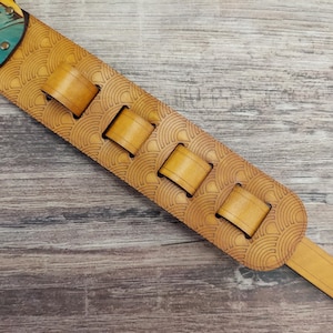 Puede incluir: Una correa de guitarra marr&oacute;n claro con un patr&oacute;n decorativo de semic&iacute;rculos. La correa tiene cuatro ranuras rectangulares y un dise&ntilde;o turquesa y amarillo en la parte superior. La correa es de cuero.