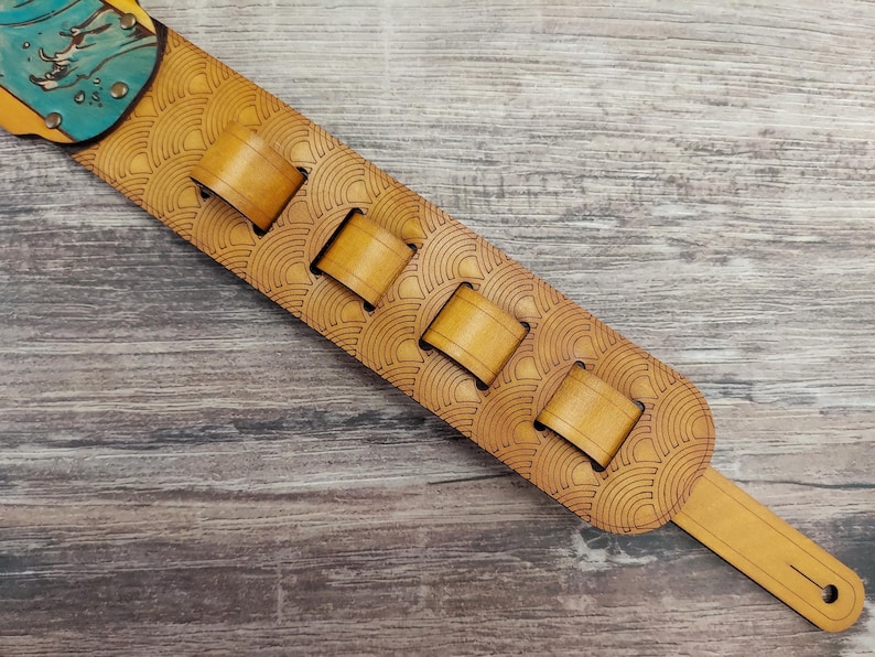 Puede incluir: Una correa de guitarra marr&oacute;n claro con un patr&oacute;n decorativo de semic&iacute;rculos. La correa tiene cuatro ranuras rectangulares y un dise&ntilde;o turquesa y amarillo en la parte superior. La correa es de cuero.