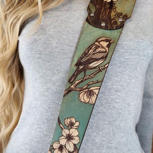 May include: A teal guitar strap with a detailed wood-burned design. The strap features a bird perched on a branch with flowers. The top portion has a tree and flower design. The strap is made of leather and has metal rivets.