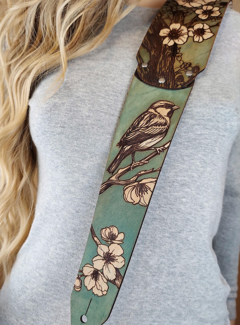 May include: A teal guitar strap with a detailed wood-burned design. The strap features a bird perched on a branch with flowers. The top portion has a tree and flower design. The strap is made of leather and has metal rivets.