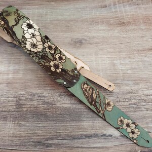May include: A guitar strap with a nature-inspired design. The strap features a teal background with a floral and bird illustration in brown and white. The strap is made of leather and has a natural wood color.