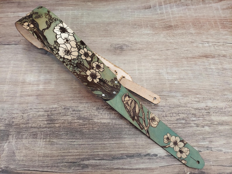 May include: A guitar strap with a nature-inspired design. The strap features a teal background with a floral and bird illustration in brown and white. The strap is made of leather and has a natural wood color.