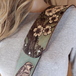 May include: A leather guitar strap with a nature-inspired design. The strap features a light blue background with a tree and flower pattern in brown and white. A detailed bird illustration is also visible.
