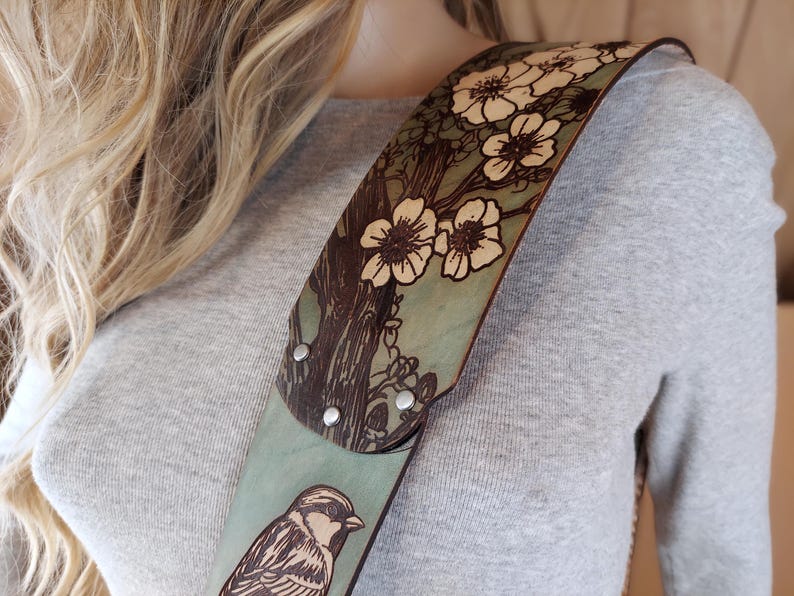 May include: A leather guitar strap with a nature-inspired design. The strap features a light blue background with a tree and flower pattern in brown and white. A detailed bird illustration is also visible.