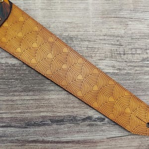Puede incluir: Una correa de guitarra con un patr&oacute;n de abanico repetido grabado en el cuero marr&oacute;n claro. La correa tiene un extremo marr&oacute;n oscuro con remaches de lat&oacute;n y un acento turquesa y amarillo.