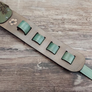 May include: A guitar strap made of light brown leather with green accents. The strap features a floral design on the shoulder pad and four green loops for adjustment. The end of the strap has a hole for attachment.