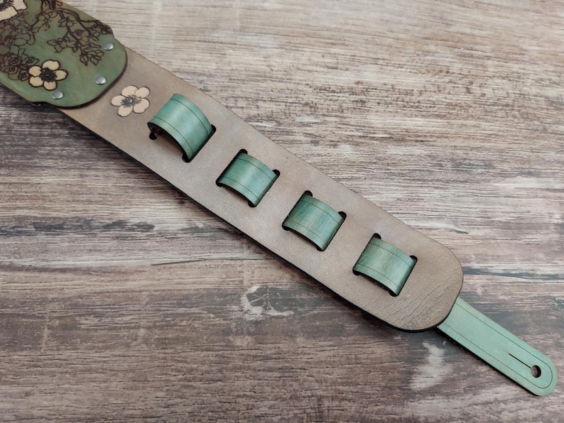 May include: A guitar strap made of light brown leather with green accents. The strap features a floral design on the shoulder pad and four green loops for adjustment. The end of the strap has a hole for attachment.