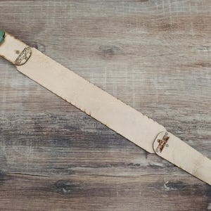 May include: A light tan leather guitar strap with a green buckle and decorative metal rivets. The strap is angled across a wooden surface, showcasing its natural texture and design.