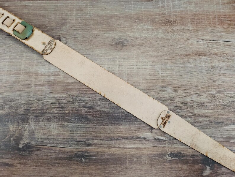 May include: A light tan leather guitar strap with a green buckle and decorative metal rivets. The strap is angled across a wooden surface, showcasing its natural texture and design.
