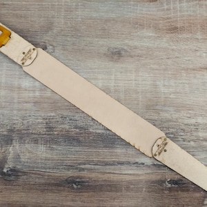 Puede incluir: Una correa de guitarra de cuero de color beige claro con un acabado natural. La correa presenta una hebilla y orificios de ajuste en un extremo y una pieza decorativa con texto en el otro. La correa se muestra sobre una superficie de madera.