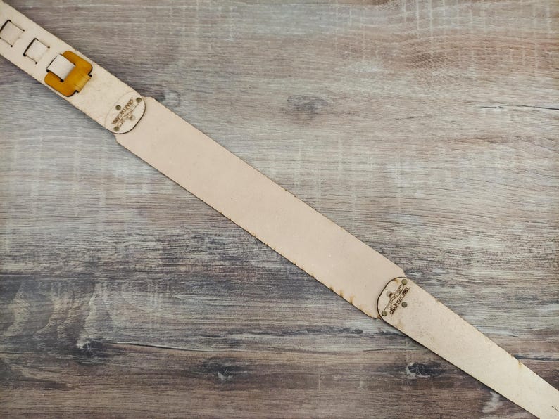 Puede incluir: Una correa de guitarra de cuero de color beige claro con un acabado natural. La correa presenta una hebilla y orificios de ajuste en un extremo y una pieza decorativa con texto en el otro. La correa se muestra sobre una superficie de madera.