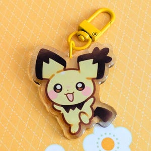 May include: A yellow and black cartoon character keychain with a yellow clasp. The character has a smiling face and is set against a yellow background with white flower accents.