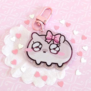 May include: A clear acrylic keychain featuring a cartoon character with a pink bow and blushing cheeks. The keychain has a pink clasp and is surrounded by pink and white heart-shaped confetti on a pink patterned background.