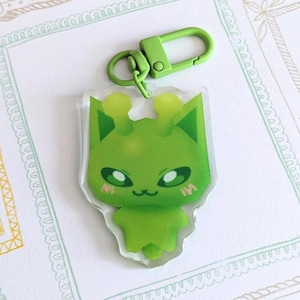 Puede incluir: Un llavero acrílico verde con un diseño de gato de dibujos animados y un cierre verde. El llavero está sobre un fondo blanco con ilustraciones de marcos decorativos en verde, amarillo y gris.