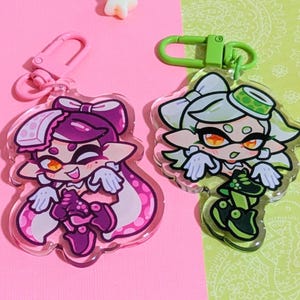 May include: Two acrylic keychains featuring cartoon characters with vibrant colors. One keychain has a pink clasp and a character with purple hair and clothing. The other has a green clasp and a character with white and green hair and clothing. Star-shaped decorations are scattered around.