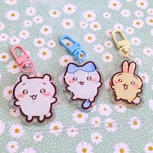 May include: Three acrylic keychains featuring cute cartoon animals: a bear, a cat, and a bunny. Each keychain has a different color clasp: pink, blue, and gold. The keychains are on a light green background with a daisy pattern. The text on the image says "These products are drawn by and sold by LittleParadeShop through Etsy. DO NOT purchase from any other source. AliExpress TikTok etc."