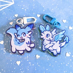 May include: Two acrylic keychains featuring cartoon-style creatures in shades of blue and white. One keychain has a white clasp, and the other has a blue clasp. The background is light blue with white geometric shapes and heart-shaped confetti.