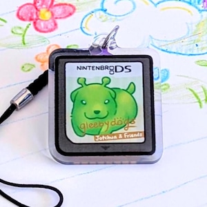 May include: A Nintendo DS game cartridge for "Sleepy Dogs" featuring a green cartoon dog. The cartridge is clear with a black lanyard attached. The background is a notebook page with colorful drawings of a rainbow, flowers, and shapes.