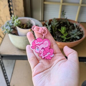 May include: A clear acrylic keychain featuring a cartoon pony character with bright pink fur, a curly pink mane, and large blue eyes. The character is winking and smiling. The keychain is attached to a light pink plastic lobster clasp hook.
