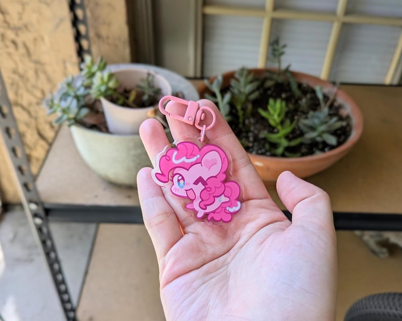 May include: A clear acrylic keychain featuring a cartoon pony character with bright pink fur, a curly pink mane, and large blue eyes. The character is winking and smiling. The keychain is attached to a light pink plastic lobster clasp hook.