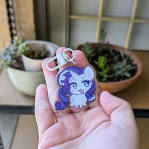 May include: A clear acrylic keychain featuring a cartoon pony character with a white body, a purple mane and tail, and a blue diamond-shaped cutie mark. The character has large blue eyes and a small horn. It includes a white plastic lobster clasp.