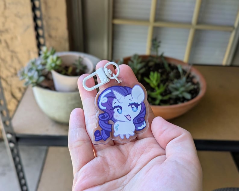 May include: A clear acrylic keychain featuring a cartoon pony character with a white body, a purple mane and tail, and a blue diamond-shaped cutie mark. The character has large blue eyes and a small horn. It includes a white plastic lobster clasp.