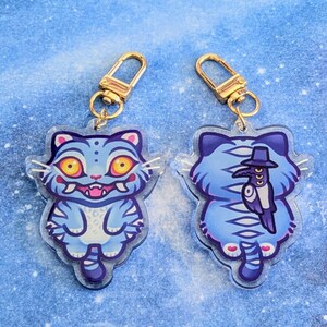 May include: Two cat-shaped keychains. The front of the cat is light blue with yellow eyes and a pink mouth. The back of the cat is light blue with a black hat and a bird design. Each keychain has a gold-colored clasp.
