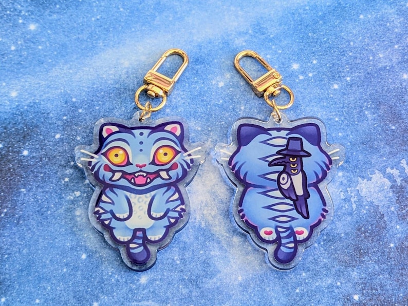 May include: Two cat-shaped keychains. The front of the cat is light blue with yellow eyes and a pink mouth. The back of the cat is light blue with a black hat and a bird design. Each keychain has a gold-colored clasp.