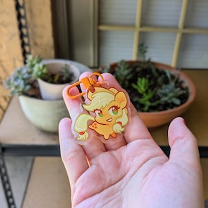 May include: A clear acrylic keychain featuring a cartoon pony character with a light orange body, blonde mane and tail, and green eyes. The pony has a small apple symbol on its flank and is attached to a bright orange plastic lobster claw clasp.