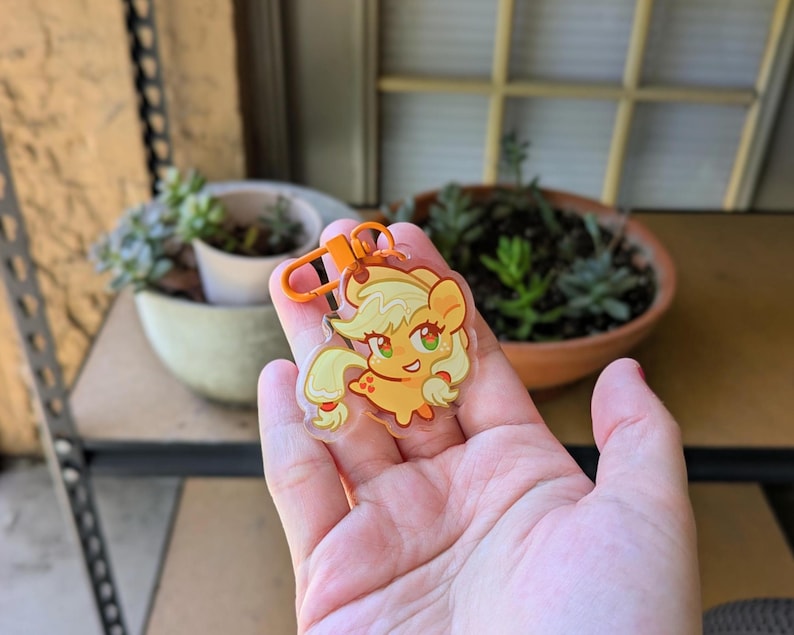 May include: A clear acrylic keychain featuring a cartoon pony character with a light orange body, blonde mane and tail, and green eyes. The pony has a small apple symbol on its flank and is attached to a bright orange plastic lobster claw clasp.