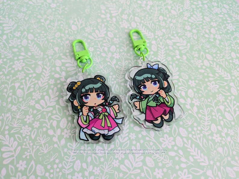 K&ouml;nnte beinhalten: Zwei transparente Acryl-Schl&uuml;sselanh&auml;nger mit Anime-Illustrationen eines M&auml;dchens in traditioneller koreanischer Hanbok-Kleidung. Die Schl&uuml;sselanh&auml;nger haben gr&uuml;ne Verschl&uuml;sse und befinden sich auf einem hellgr&uuml;nen floralen Hintergrund. Das Outfit des M&auml;dchens ist rosa und gr&uuml;n.