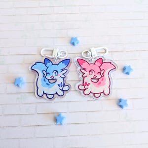 May include: Two acrylic keychains featuring cartoon-style creatures, one blue and one pink, each with a silver clasp. The blue keychain has white accents, and the pink one has red accents. Small blue star-shaped decorations are scattered around the keychains.