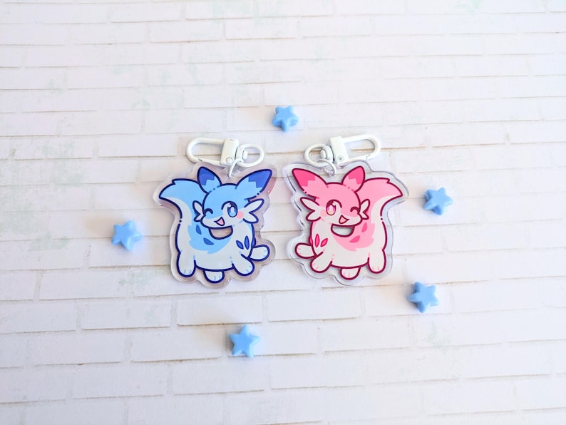 May include: Two acrylic keychains featuring cartoon-style creatures, one blue and one pink, each with a silver clasp. The blue keychain has white accents, and the pink one has red accents. Small blue star-shaped decorations are scattered around the keychains.