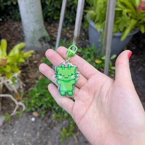 May include: A green alien cat keychain with a clear plastic background. The keychain is attached to a silver metal ring.