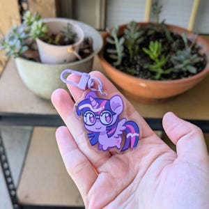 May include: A clear acrylic keychain featuring a cartoon pony character with purple fur, a dark purple mane with pink stripes, and large glasses. The character has a small horn and wings, and is attached to a light purple plastic lobster clasp hook.