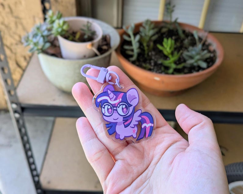 May include: A clear acrylic keychain featuring a cartoon pony character with purple fur, a dark purple mane with pink stripes, and large glasses. The character has a small horn and wings, and is attached to a light purple plastic lobster clasp hook.